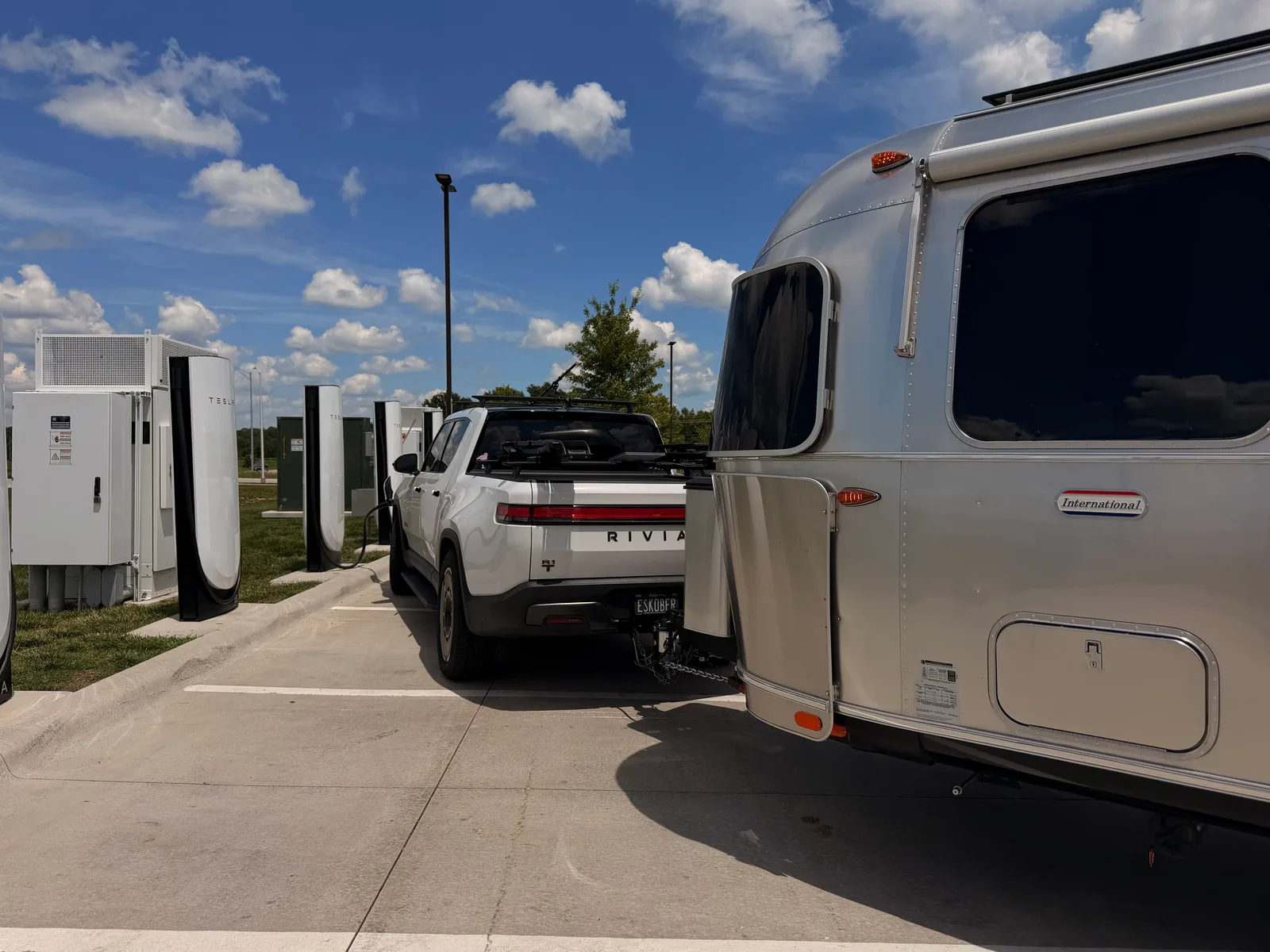 The white Rivian R1T and silver Airstream parked at the Tesla Supercharger in Springfield, MO, red-and-white stalls under a bright sky