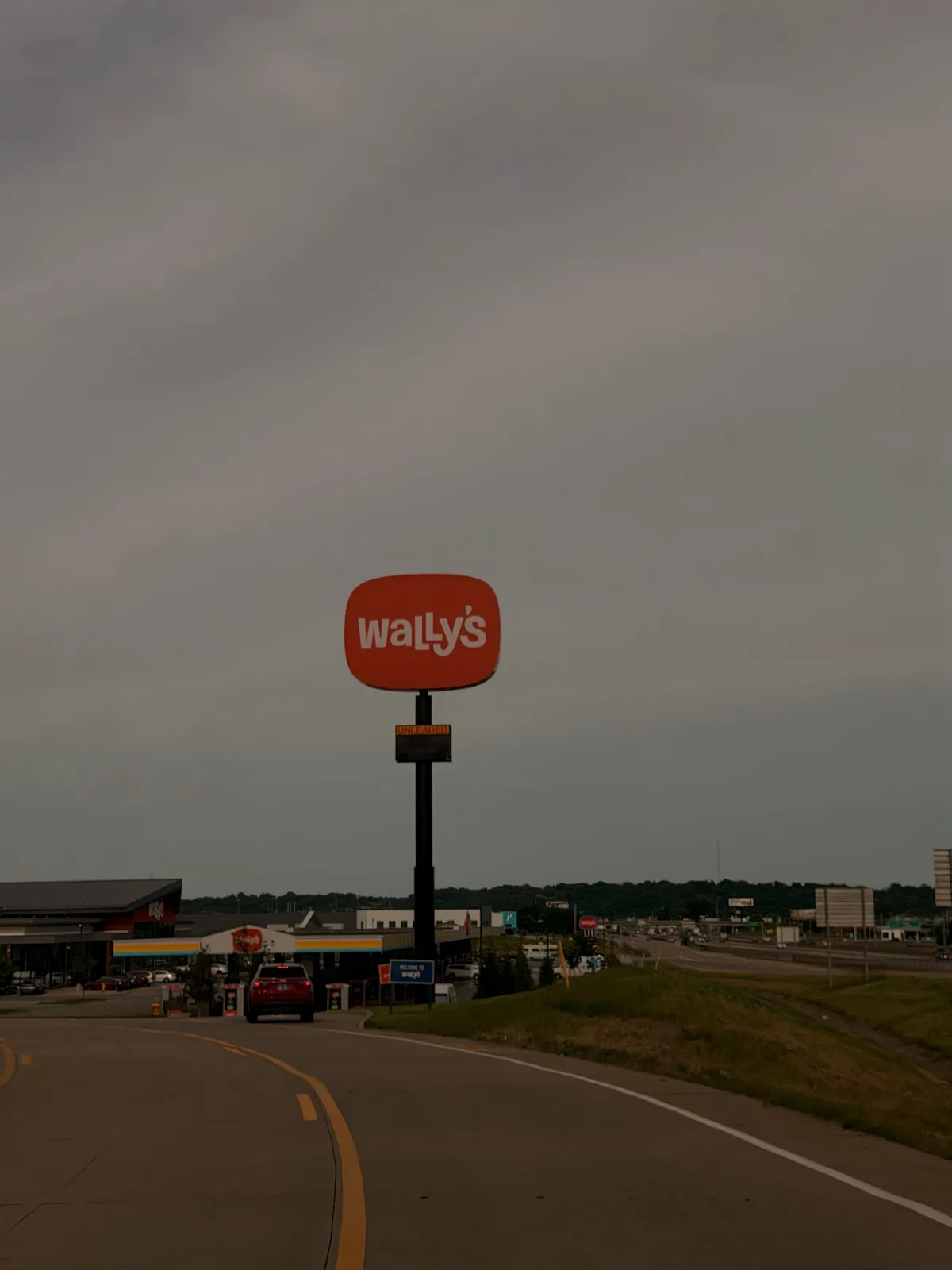 The tall illuminated orange Wally's travel-center sign rising over the Fenton MO interchange under cloudy skies, the fuel canopy below
