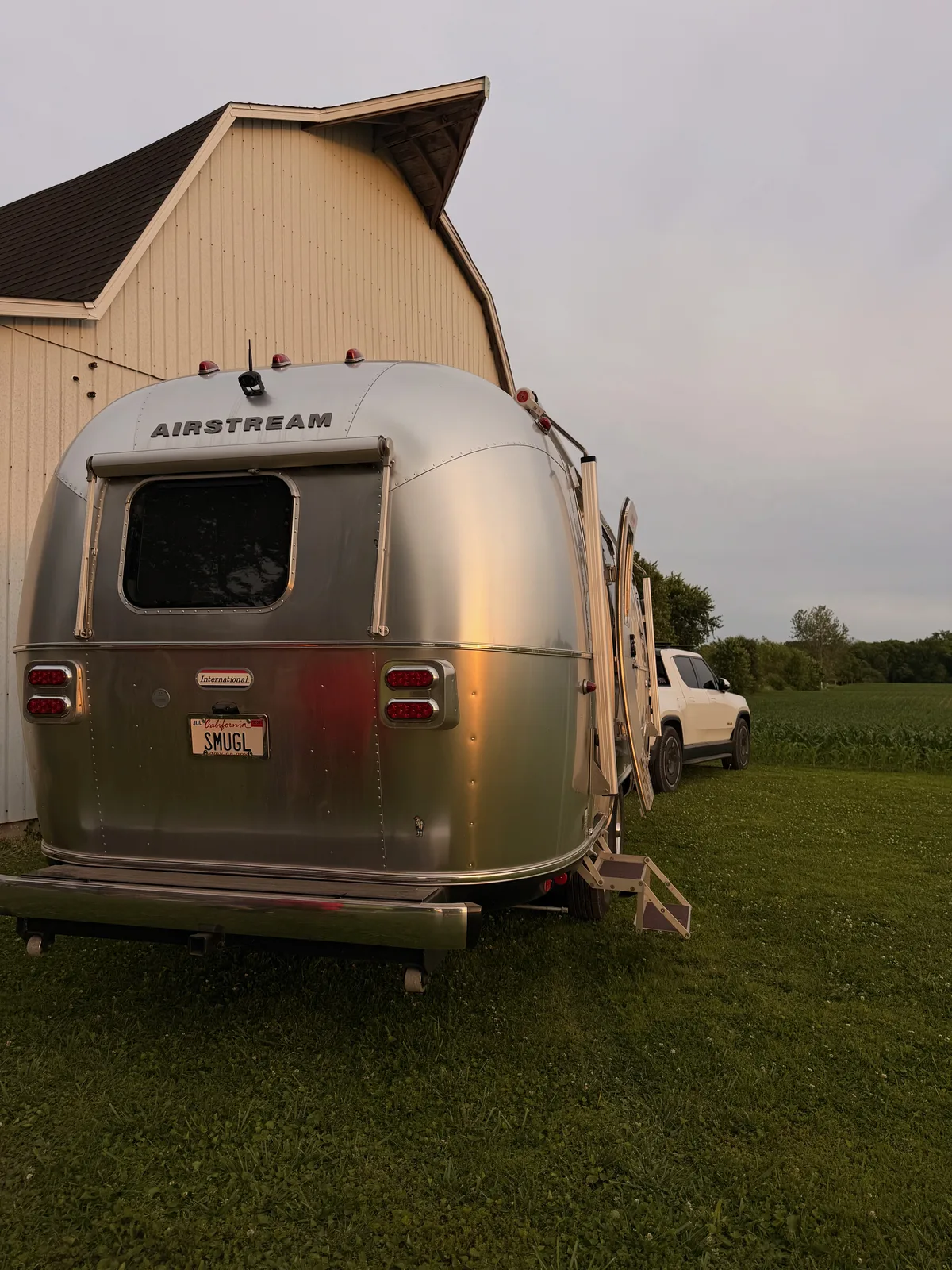 The silver Airstream parked on a grassy farm pad at dusk beside a pickup truck, an open Illinois field and tree line behind