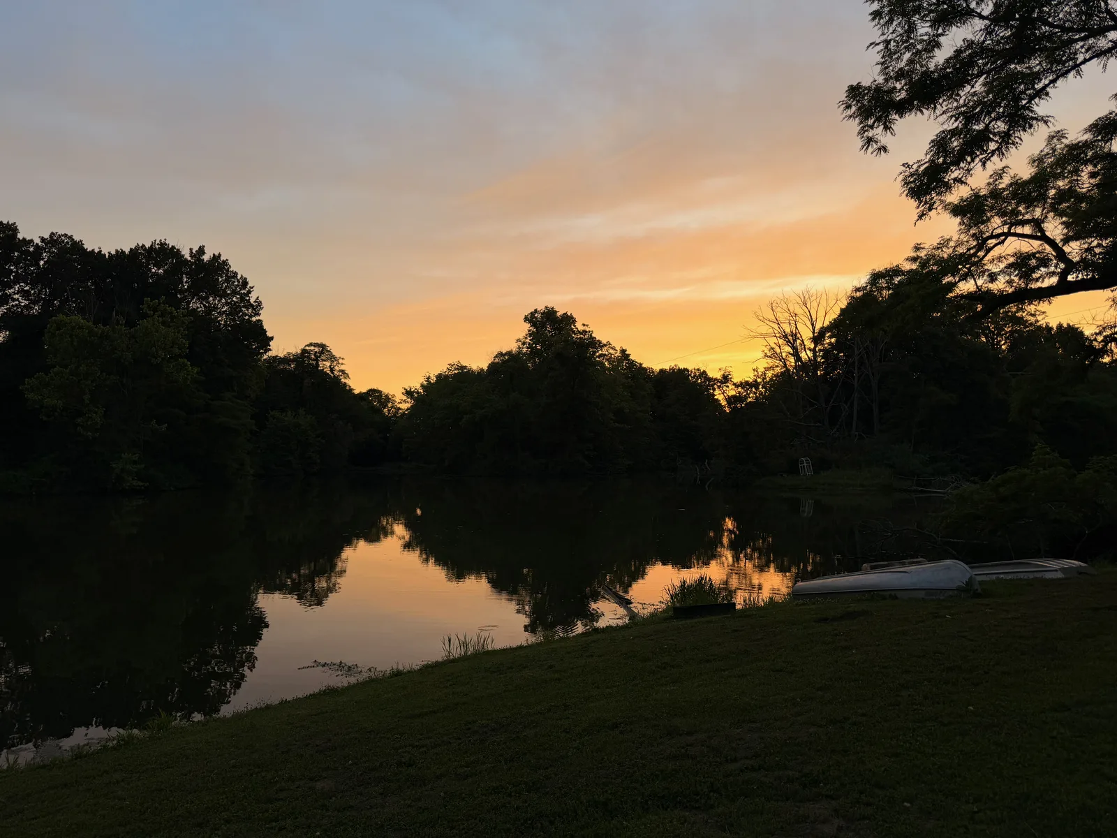 A still farm pond at sunset with an orange and gold sky mirrored on the water, a tree line and a canoe along the far bank