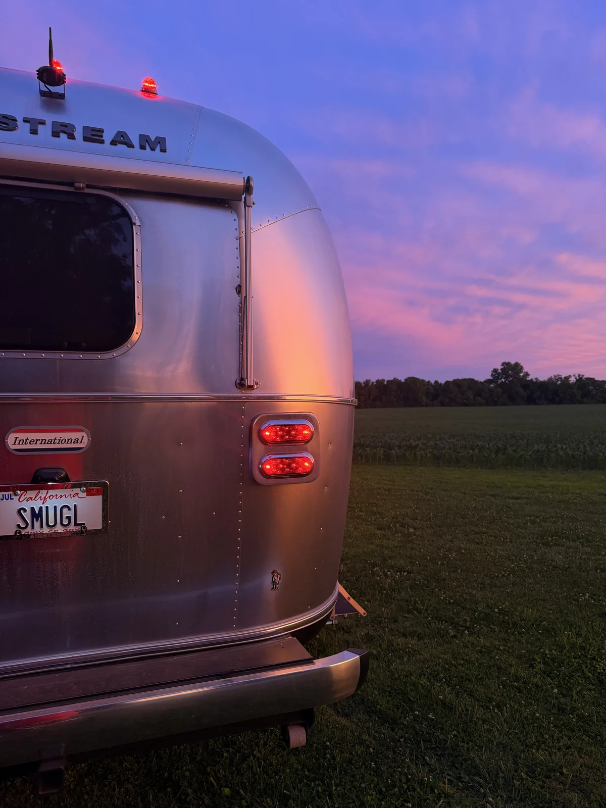 The rear of the silver Airstream against a purple and pink sunset at the Illinois farm, the SMUGL vanity plate reading Smugglers Escape