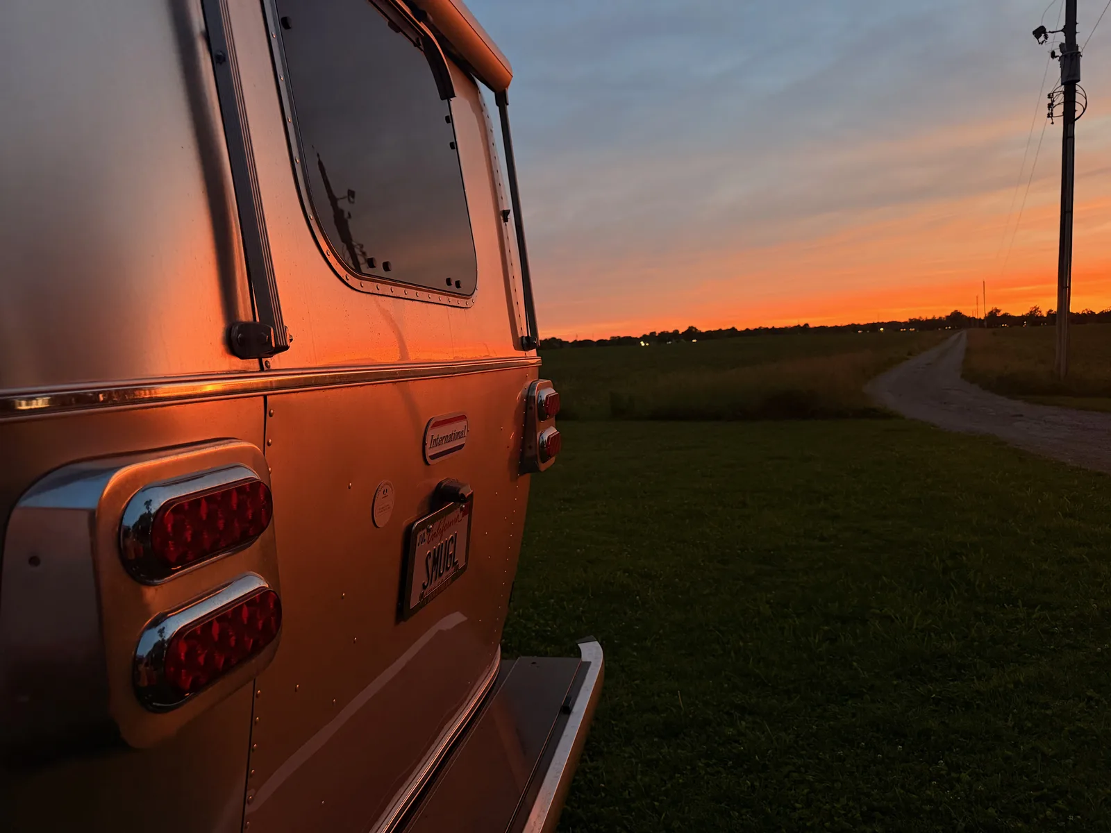 The rear of the silver Airstream at sunset on the farm, a gravel drive curving out across the open field under an orange sky