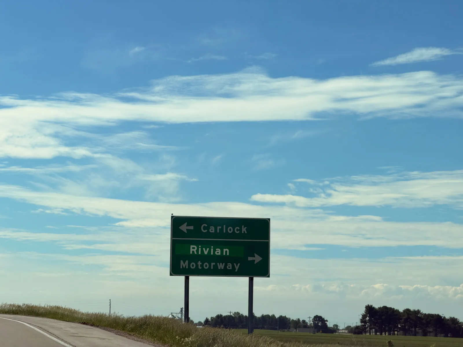 A green highway sign near Carlock, Illinois pointing left to Carlock and right to Rivian Motorway, the road to Rivian's Normal plant, under a clear blue sky