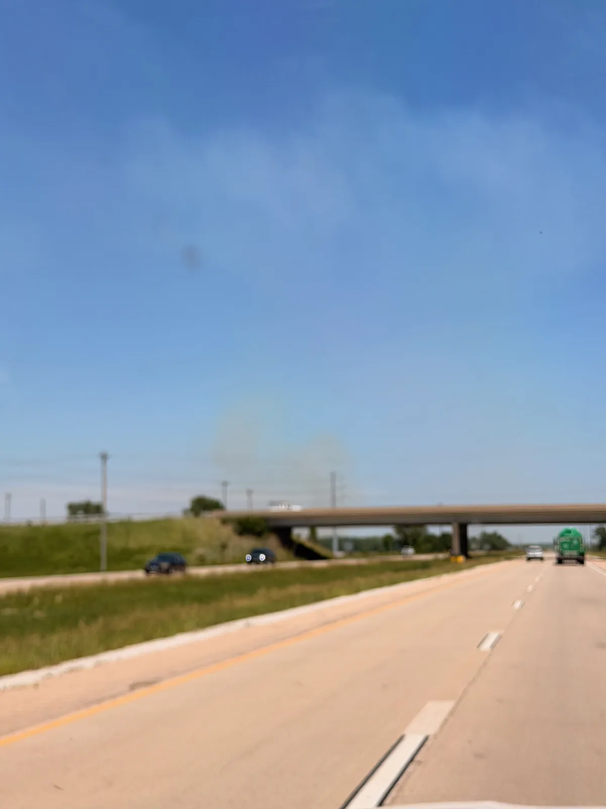 A column of dark smoke rising over the empty I-39 horizon from a distant burn, the highway running ahead under blue sky