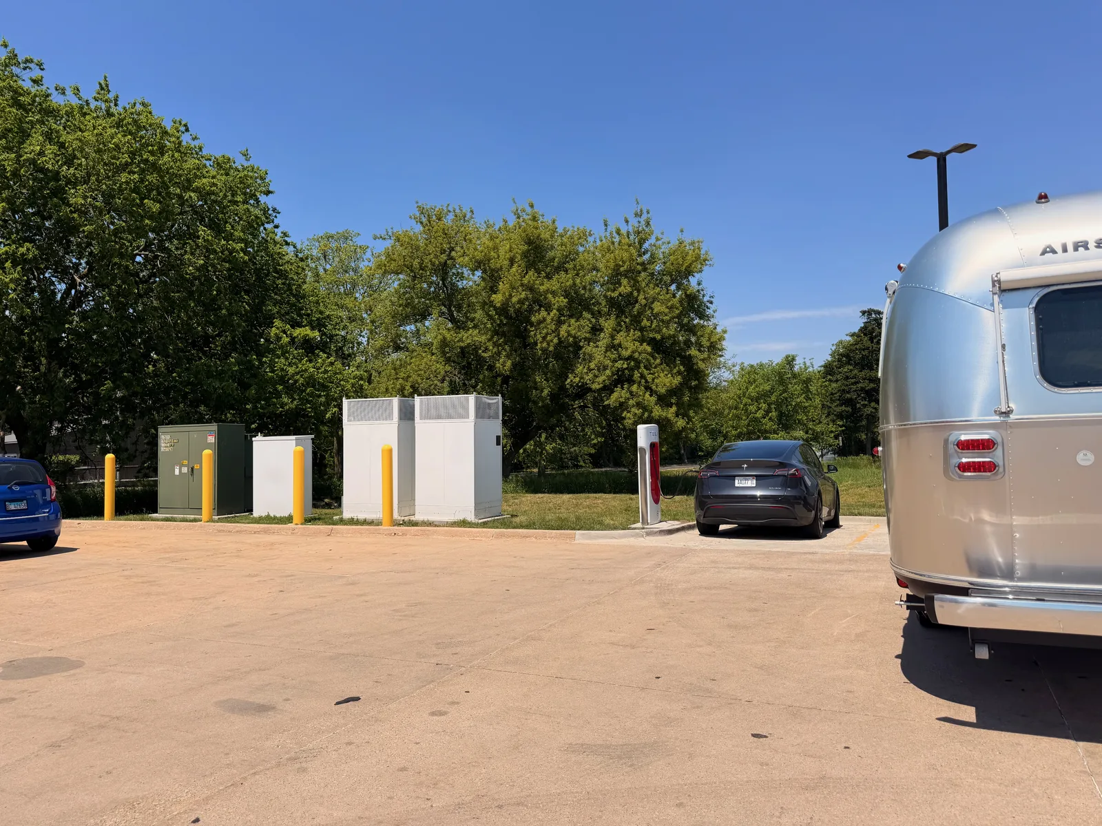 The white Rivian R1T and the silver Airstream parked at a row of tall white Tesla Supercharger pedestals at the Rochelle, Illinois site under a clear blue sky, the day's first third-party SC attempt