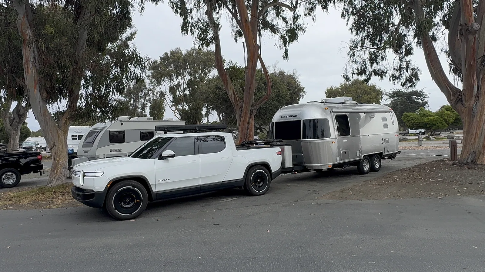 The white Rivian R1T and the silver Airstream parked together at a wooded roadside pullout, sunlight filtering through the trees