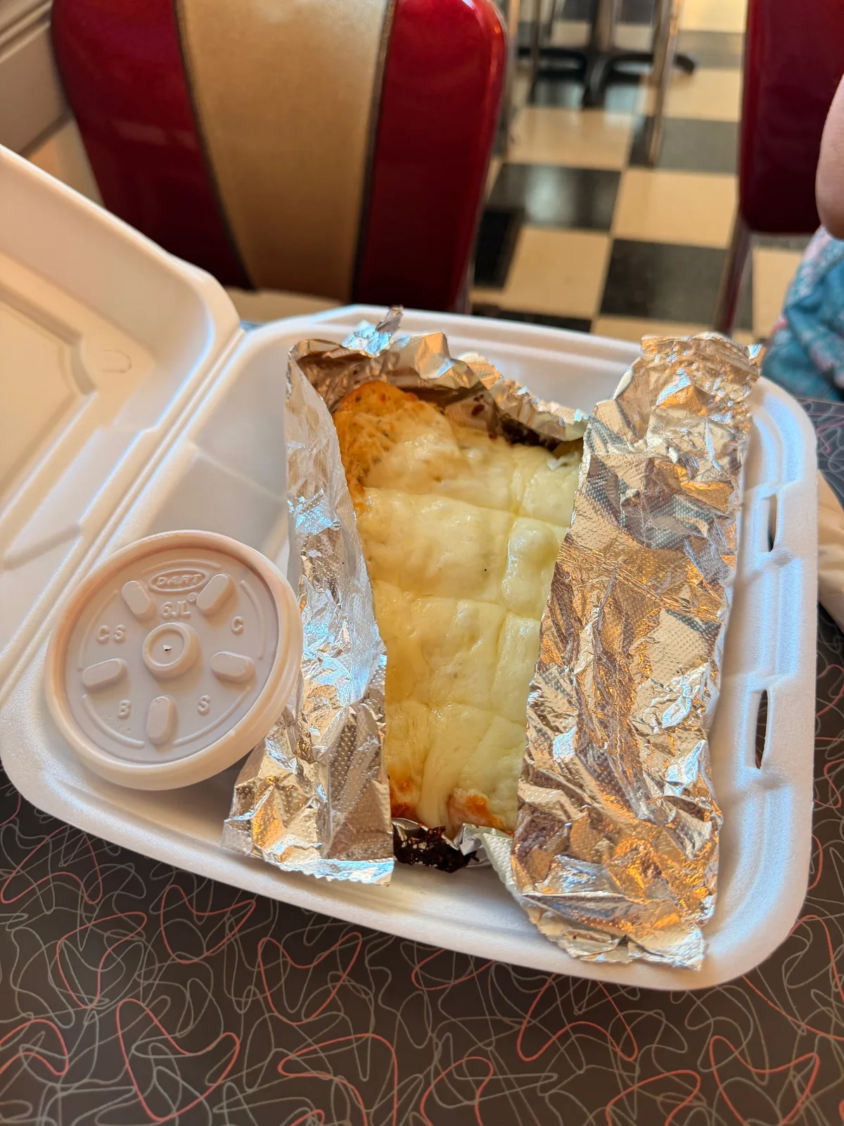 An Italian beef sandwich in a foil-lined foam takeout box on a diner table at Orlando's in Round Lake, a red vinyl booth and black-and-white checkered floor behind it