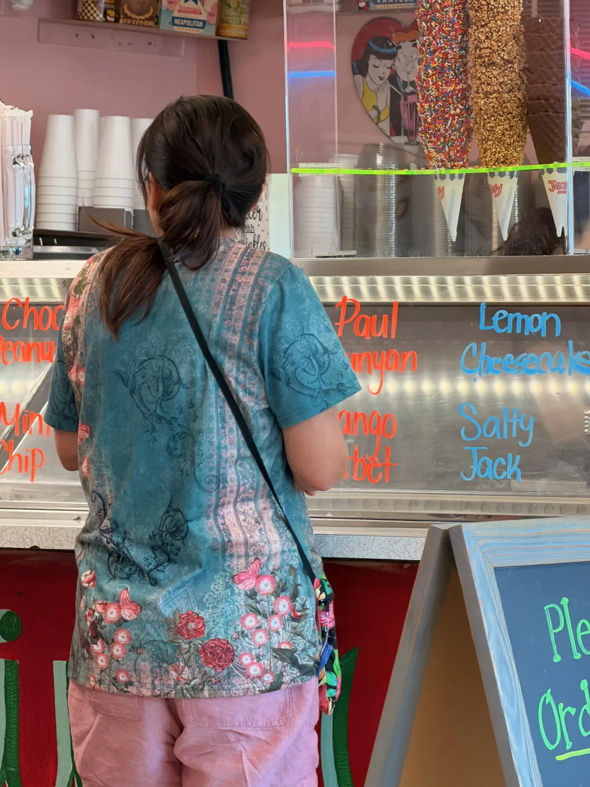 The flavor board behind the counter glass at Diana's Ice Cream Shop in Round Lake listing Paul Bunyan, Mango Sorbet, Lemon Cheesecake, and Salty Jack, a customer ordering at the window
