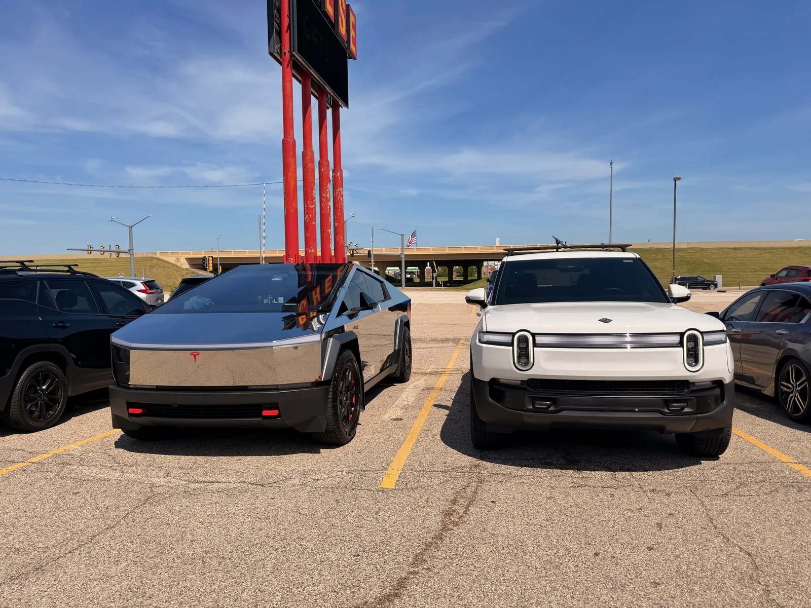 The white Rivian R1T parked nose to nose with a stainless Tesla Cybertruck in the Mars Cheese Castle lot in Kenosha, the tall red Mars Cheese Castle sign and an I-94 overpass behind them