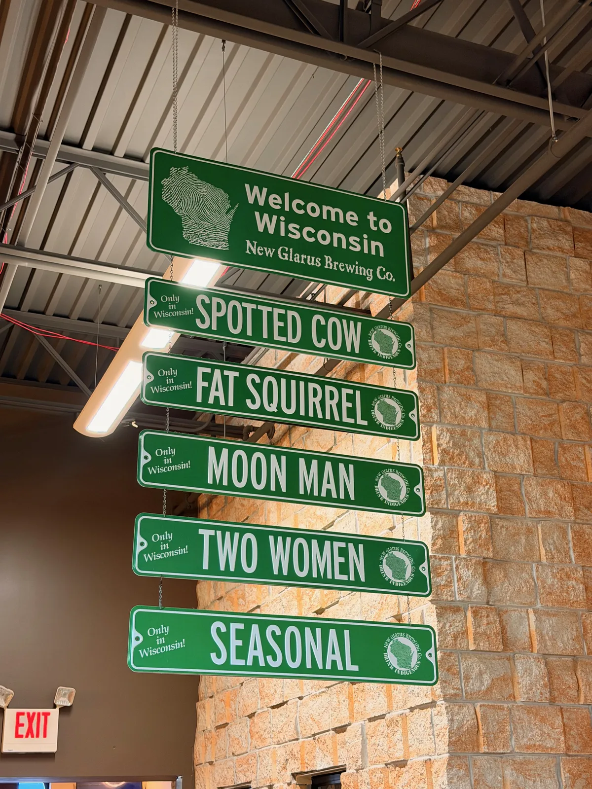 Hanging green New Glarus Brewing signs inside Mars Cheese Castle reading Welcome to Wisconsin, Spotted Cow, Fat Squirrel, Moon Man, Two Women, and Seasonal, against a stone wall