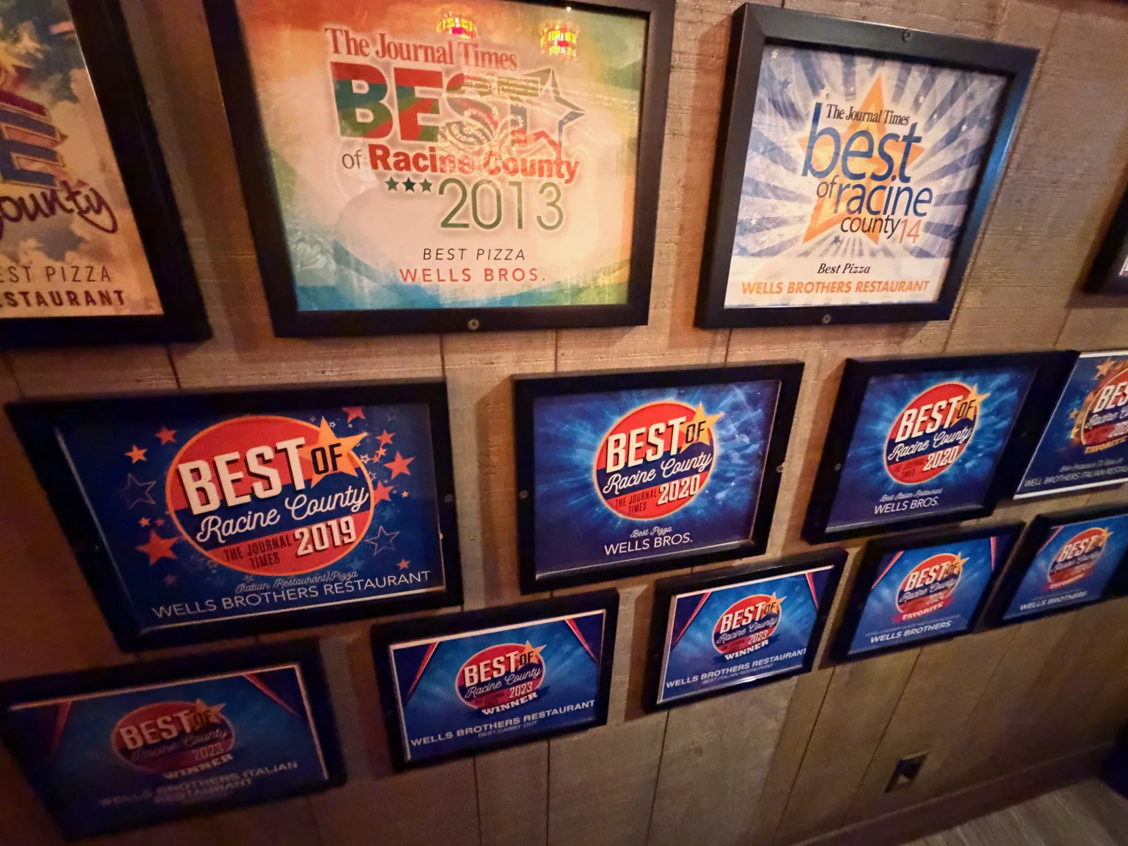 A wall of framed Best of Racine County Best Pizza awards for Wells Brothers Restaurant dating from 2013 onward, mounted on wood paneling