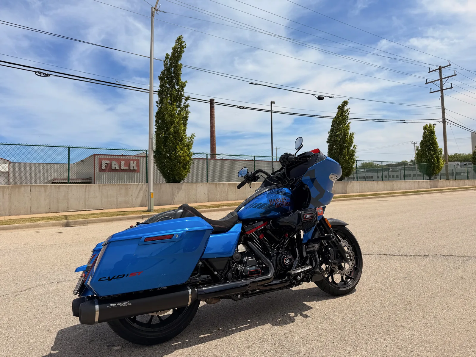 A Milwaukee-blue Harley-Davidson CVO ST Road Glide parked on the pavement in the Menomonee Valley, the old FALK works building behind it under a bright sky
