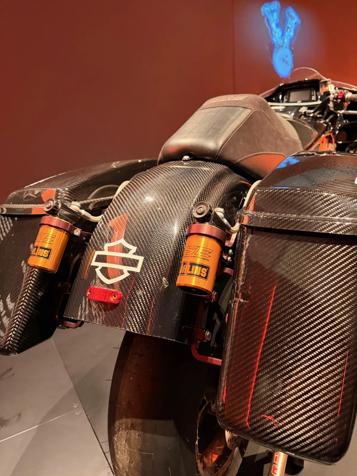 A carbon-fiber Harley-Davidson factory race bike with gold Öhlins shocks on display against a dark red wall in the Harley-Davidson Museum