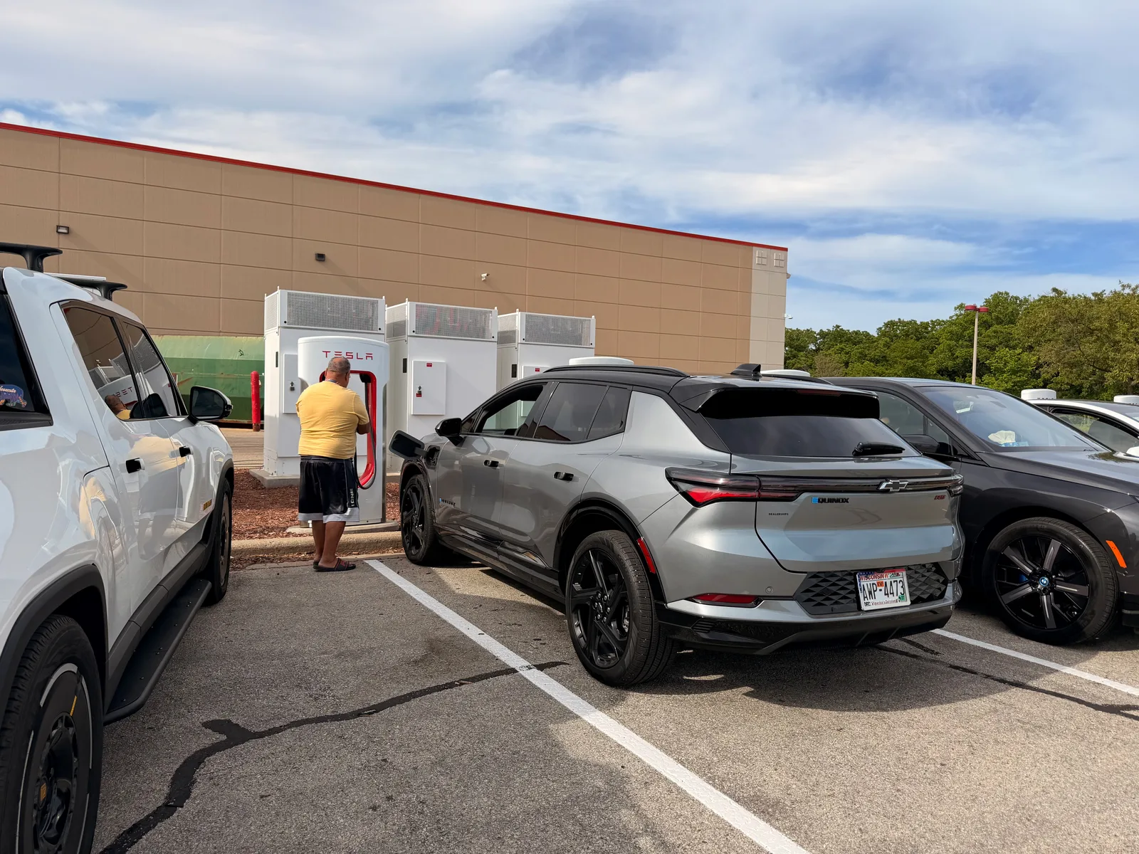 The white Rivian R1T charging at the Kenosha Tesla Supercharger next to a silver Chevrolet Equinox EV, a row of Tesla pedestals along the building