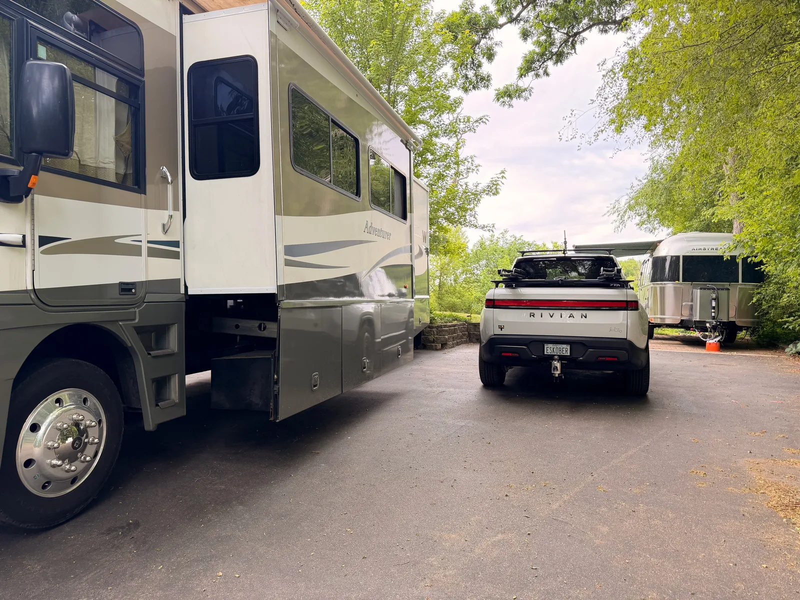 The white Rivian R1T parked under leafy trees near Fox Lake, Illinois, a polished Airstream and a motorhome alongside it