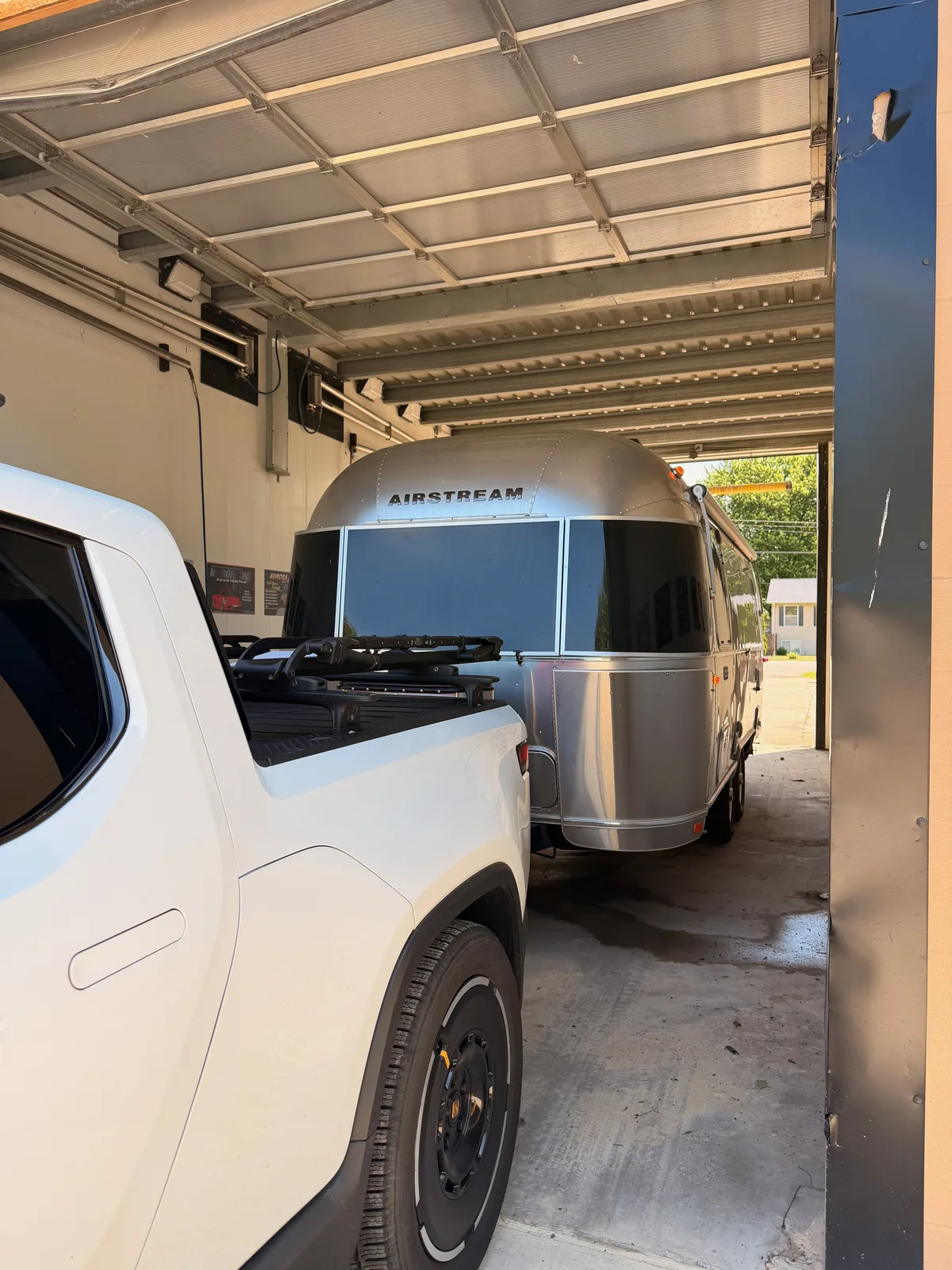 The white Rivian R1T and the polished Airstream parked together inside an open-sided car-wash bay, a gas station visible through the far opening