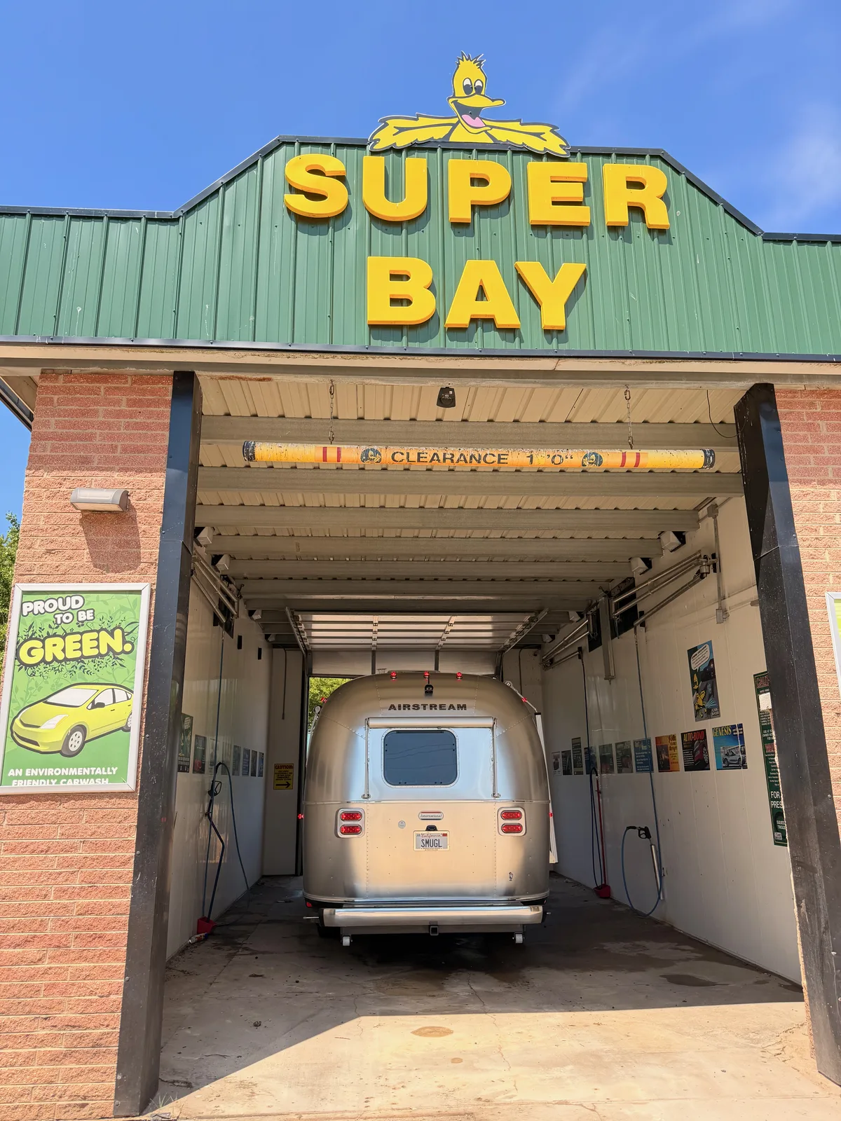 The exterior of Hub City Car Wash in Rochelle Illinois, a green-and-yellow Super Bay with a cartoon duck mascot and a 10-foot clearance sign, the Airstream parked inside the tall bay
