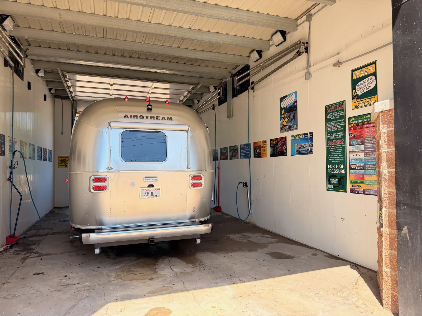 The rear of the polished aluminum Airstream parked in a self-serve high-pressure car-wash bay, its SMUGL plate visible, pressure-washer signs along the walls