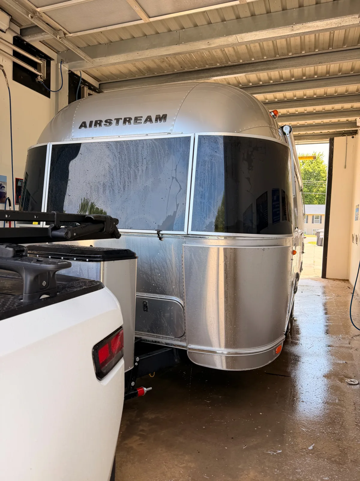 The freshly washed front of the polished aluminum Airstream gleaming under the lights of a car-wash bay, hitched to the white Rivian R1T with water pooled on the concrete floor