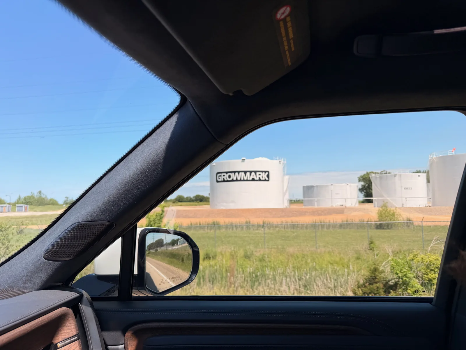 A view out the Rivian's passenger window of GROWMARK fuel-storage tanks across a fenced field under a clear blue sky, the side mirror in frame