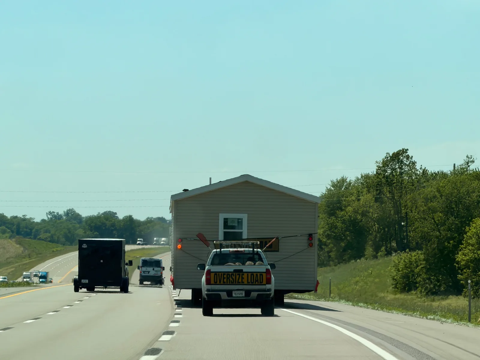 The view from the Rivian on I-80 in Iowa following a flatbed hauling an entire house, "Oversize Load" banner across the back, rolling green hills beyond