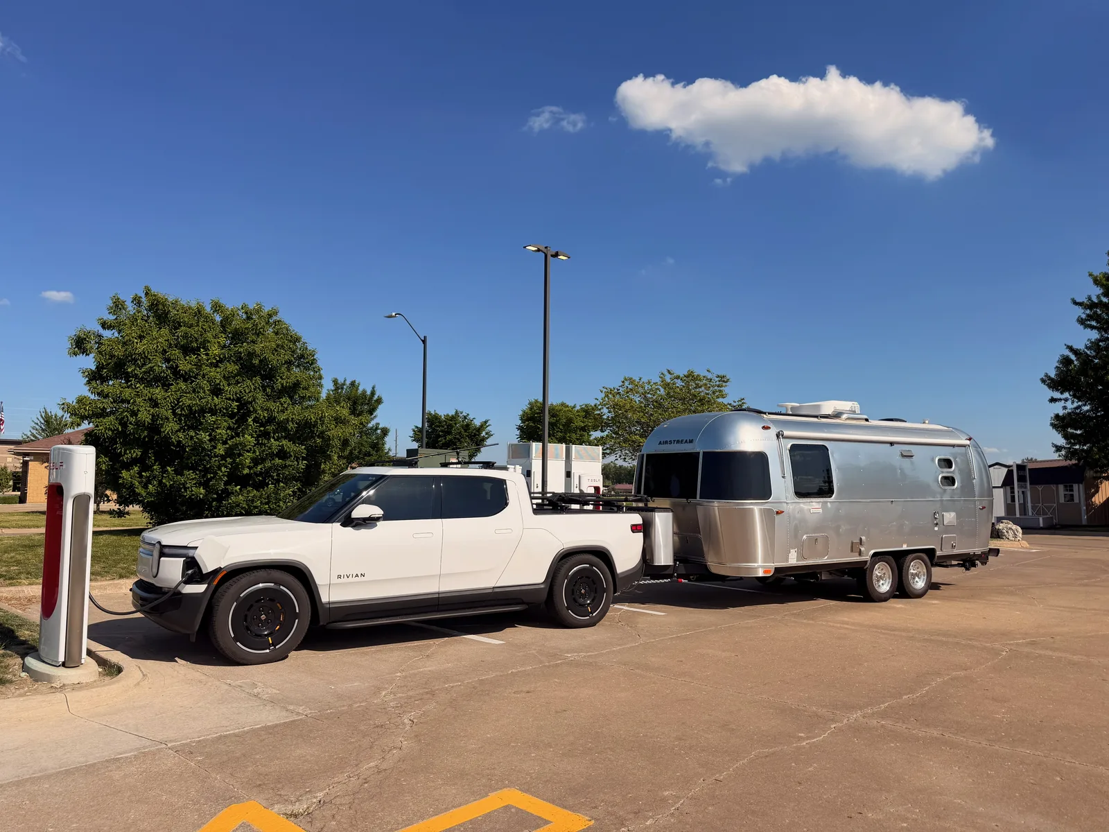 The white Rivian R1T and hitched Airstream plugged in at a Tesla Supercharger in a flat lot in Mount Pleasant, southeast Iowa, under a clear blue sky