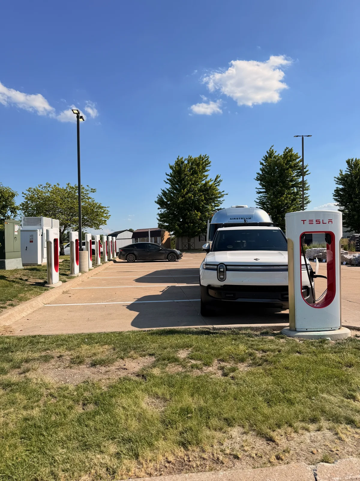 The white Rivian R1T and Airstream beside a red Tesla Supercharger post and a row of stalls, a black Tesla also charging, in Mount Pleasant Iowa