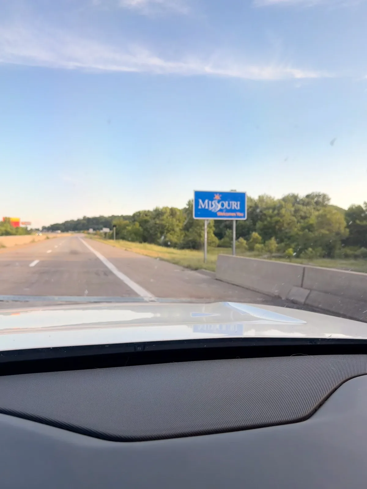 A blue "Missouri Welcomes You" state-line sign seen through the Rivian's windshield over the white hood on a clear evening, US-61 ahead