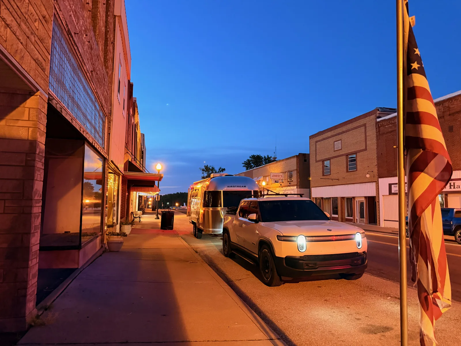 The white Rivian R1T and polished Airstream parked on a brick small-town main street at blue-hour dusk in Paris, Missouri, an American flag and warm streetlights alongside