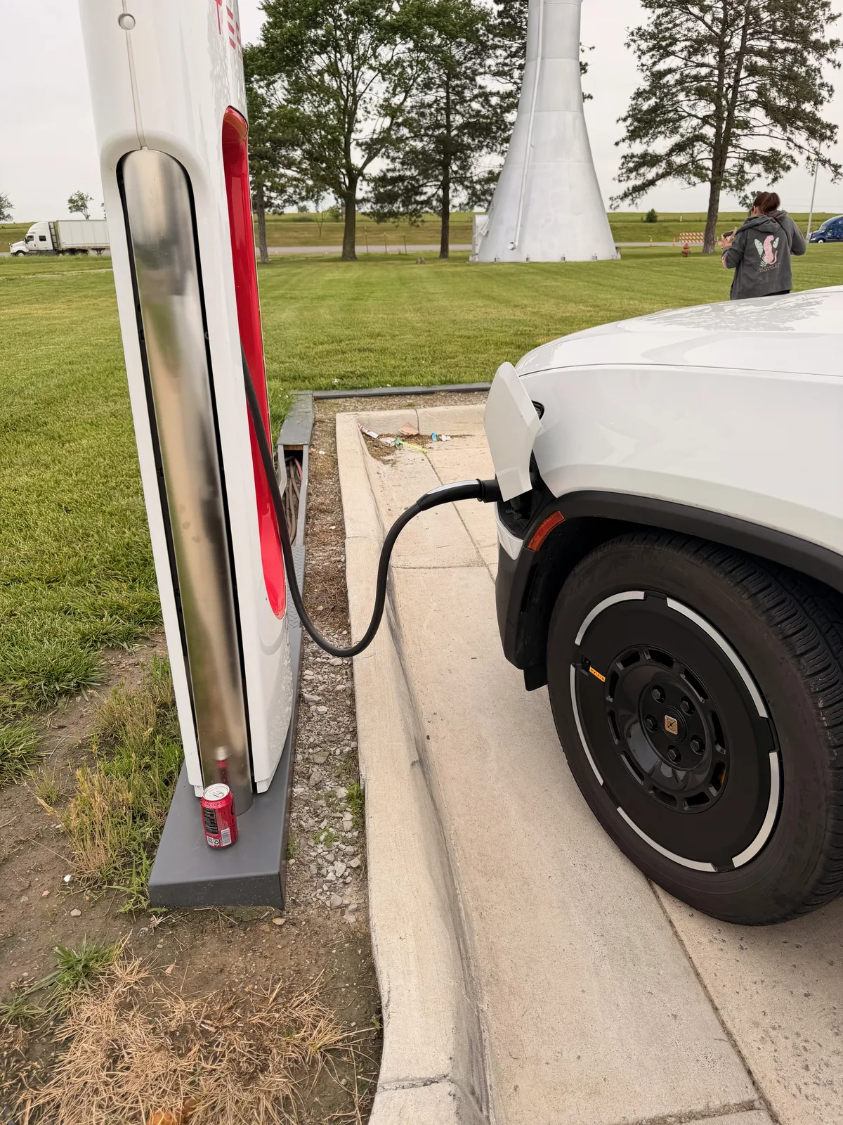 The white Rivian R1T plugged into a fast charger on a grassy lot near Topeka, wind turbines turning in the background