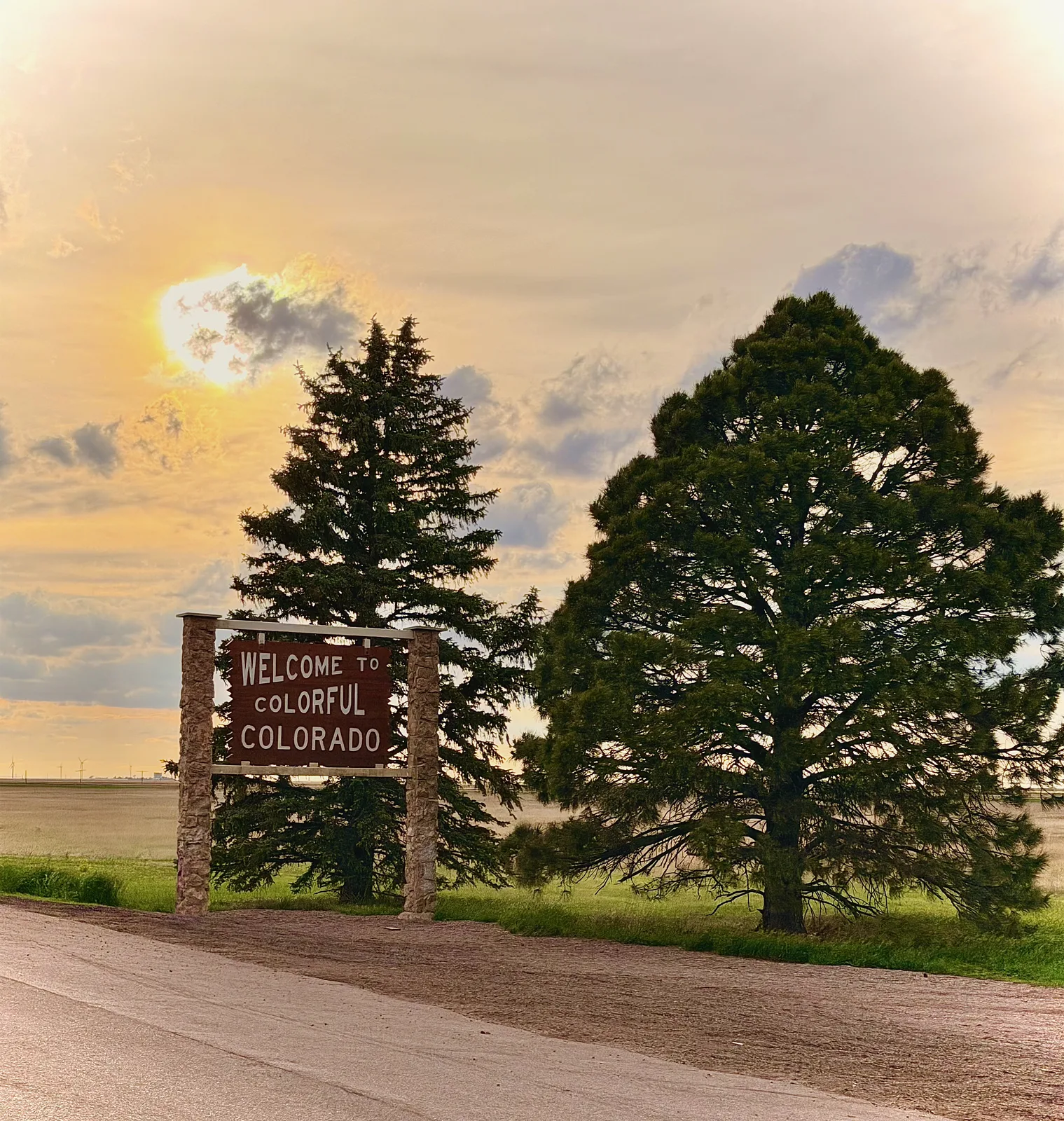 The brown and tan Welcome to Colorful Colorado sign on a stone base beside two evergreens at golden hour, the high plains and a glowing sunset sky behind it at the Kansas-Colorado line on I-70
