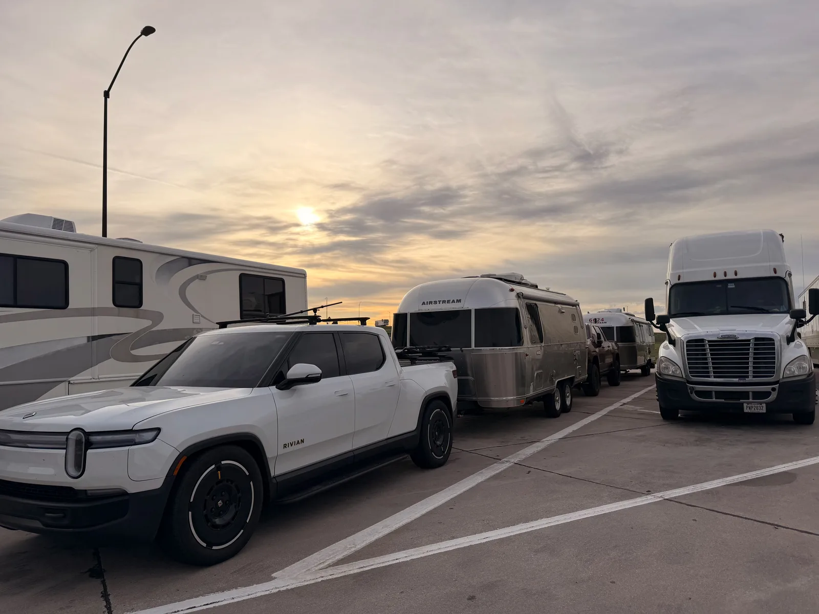 The white Rivian R1T hitched to the polished Airstream at the Arriba rest area at dawn, a Class A motorhome and a Freightliner semi alongside as the sun breaks through the clouds