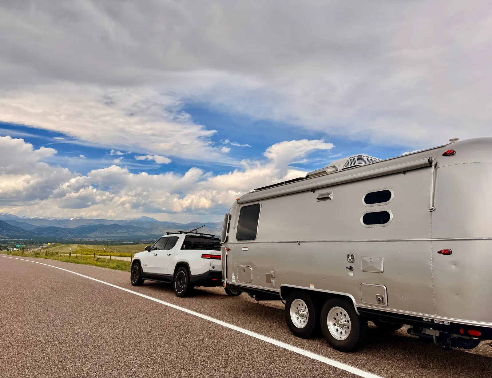 The white Rivian R1T towing the polished Airstream west on an open highway, the Front Range foothills rising on the horizon