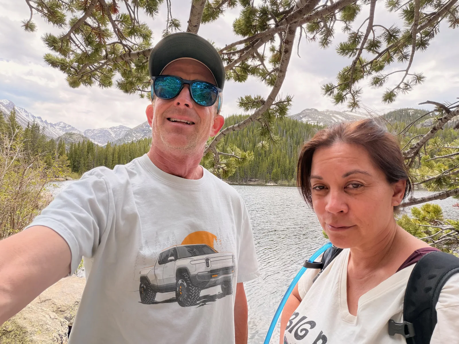 A couple taking a selfie at Bear Lake in Rocky Mountain National Park, snow-streaked peaks and pine forest across the water behind them, the man in a white Rivian R1T t-shirt