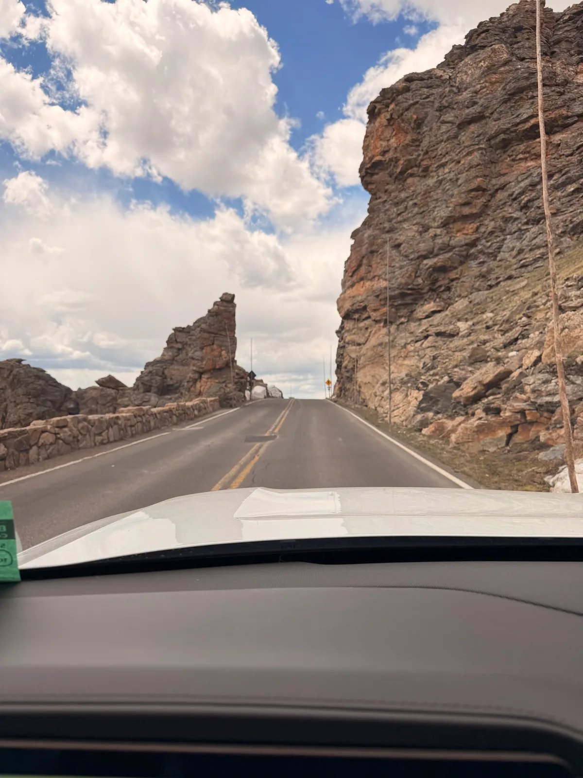 Windshield view from the white Rivian R1T on Trail Ridge Road at the Rock Cut, a jagged granite outcrop beside the two-lane road under big afternoon clouds in Rocky Mountain National Park