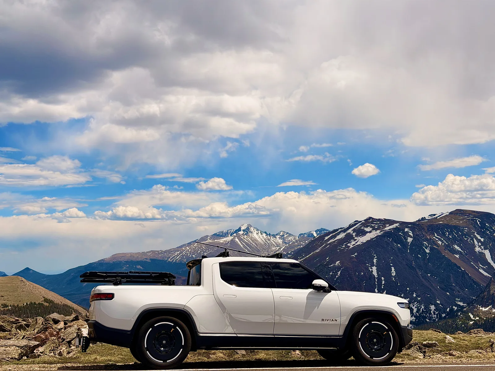 The white Rivian R1T parked in profile on Trail Ridge Road in Rocky Mountain National Park, the snow-streaked summit of Longs Peak and the Front Range behind it under towering clouds