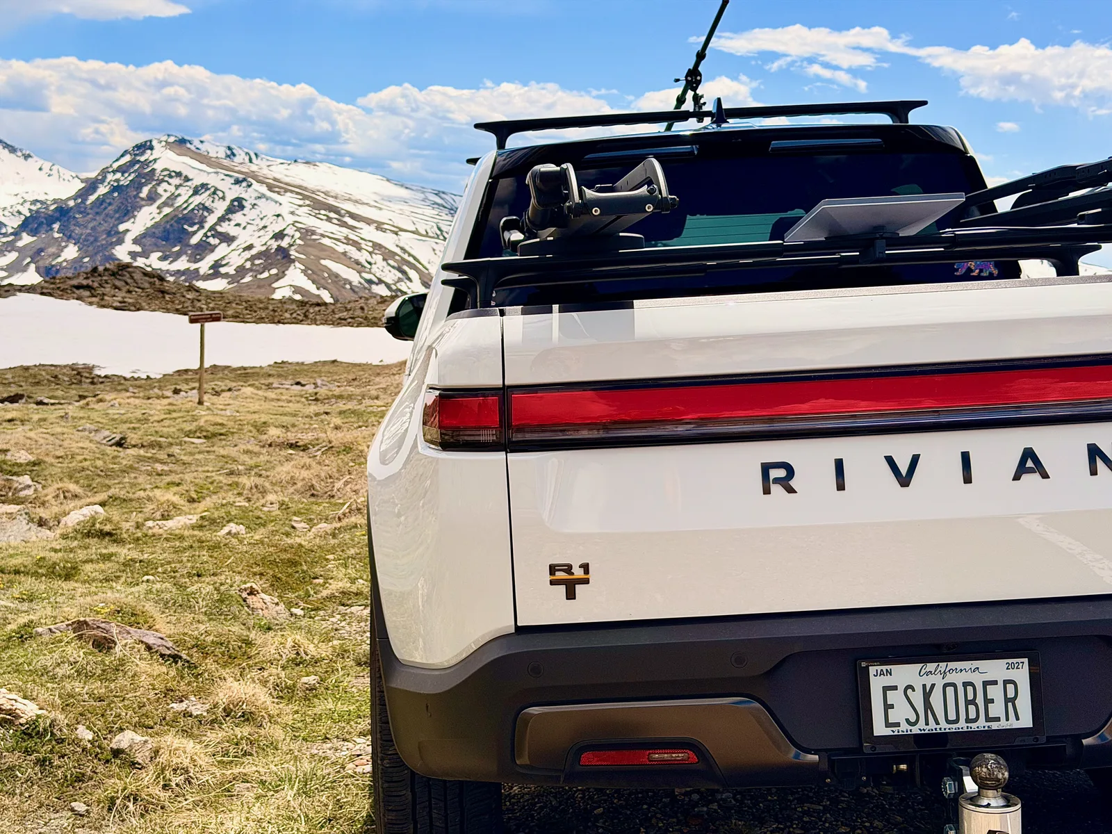 Rear view of the white Rivian R1T with its California ESKOBER plate, parked on the alpine tundra in Rocky Mountain National Park with snowfields and a snow-covered ridge behind