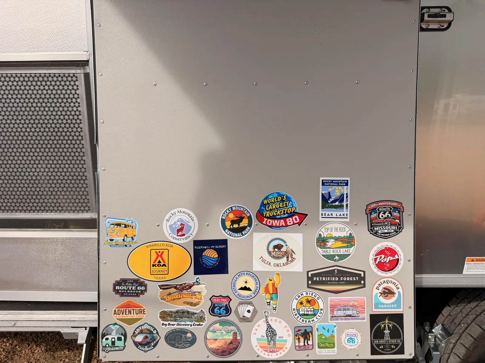 The polished Airstream's endcap covered in travel stickers including Rocky Mountain National Park, Bear Lake, Route 66, Iowa 80, and Petrified Forest, documenting the trip