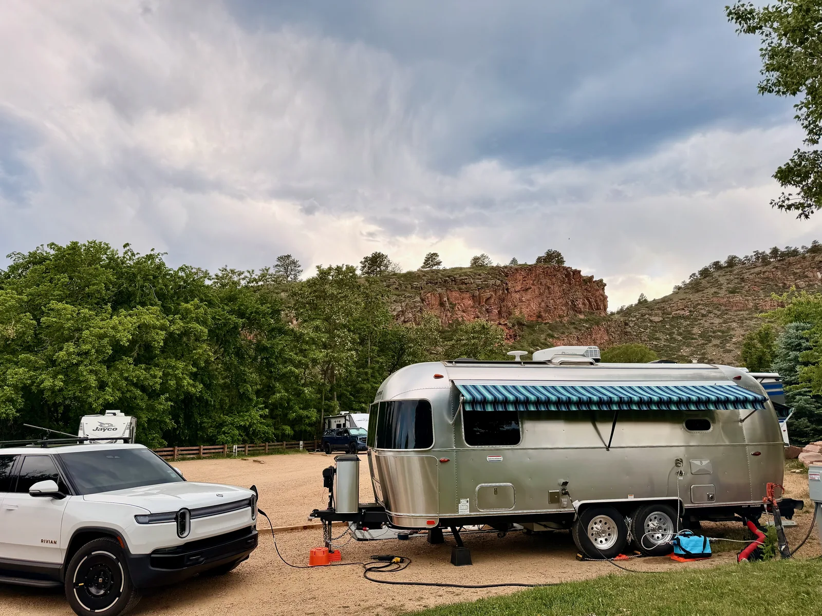 The white Rivian R1T plugged in beside the polished Airstream at the LaVern Johnson campsite in Lyons, red sandstone cliffs and a stormy evening sky behind