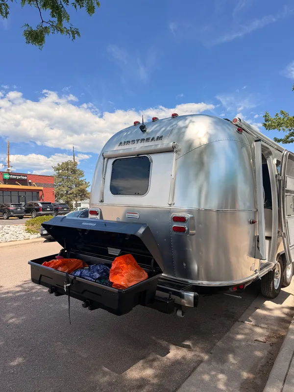 The open black Yakima cargo box on the back of the polished Airstream loaded with orange dry bags and gear, an Enterprise sign across the Rack Attack lot behind