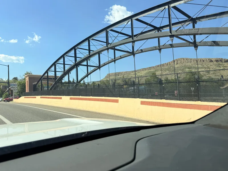 A bridge over Clear Creek in downtown Golden, Colorado seen past the white Rivian R1T's hood, the foothills rising behind under a blue sky
