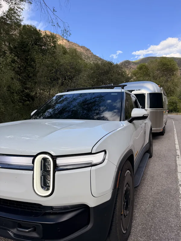The white Rivian R1T towing the polished Airstream on I-70 deep in Glenwood Canyon, sheer red-rock walls rising on both sides above the Colorado River