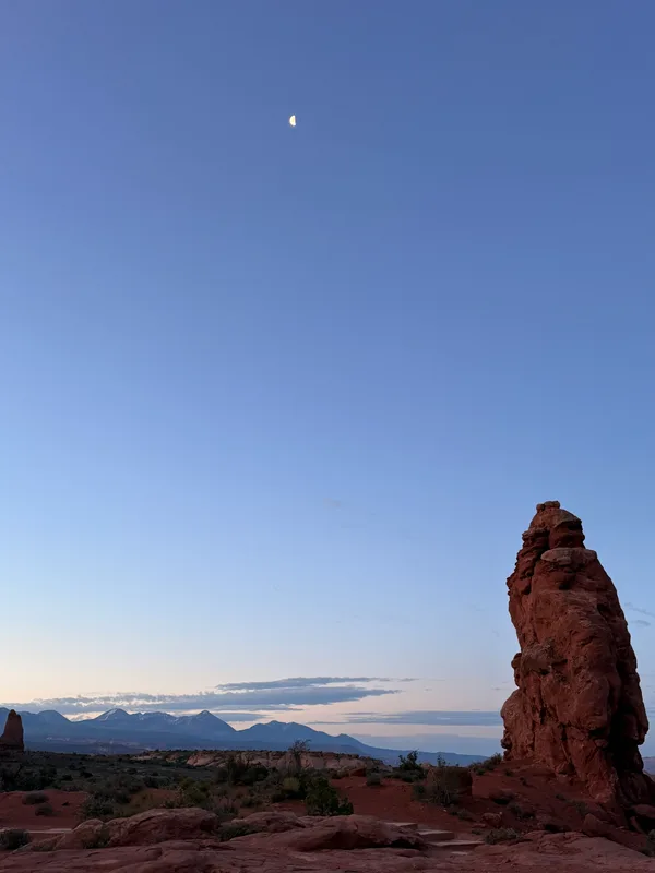 A tall red-rock spire catching the first warm light at dawn in Arches National Park, snow-dusted La Sal Mountains behind