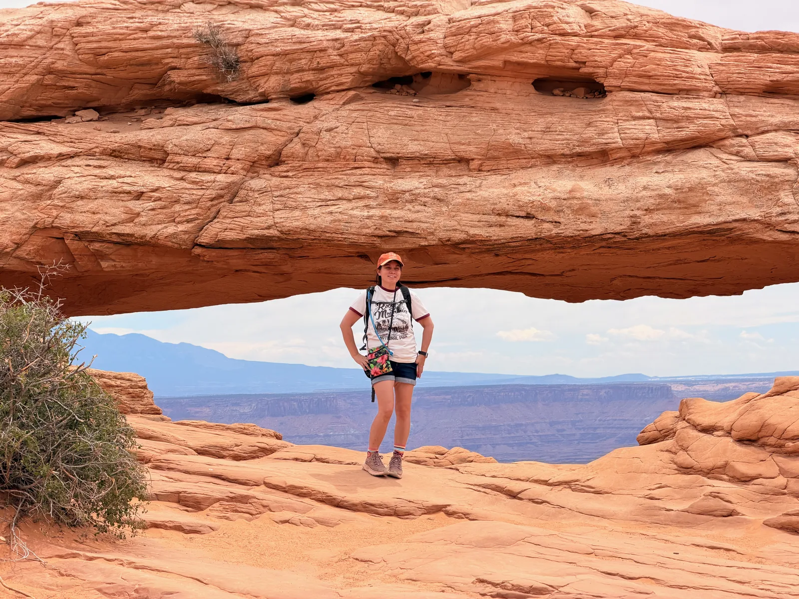 Diana standing beneath Mesa Arch in Canyonlands National Park, the desert canyon stretching out under the rock span behind her