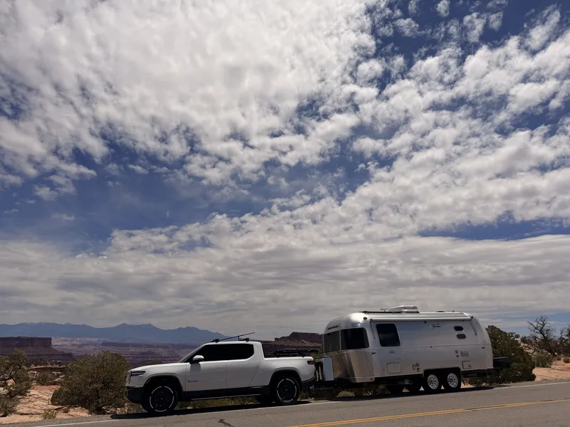 The white Rivian R1T and polished Airstream parked along the Island in the Sky road under a huge sky of scattered cumulus clouds