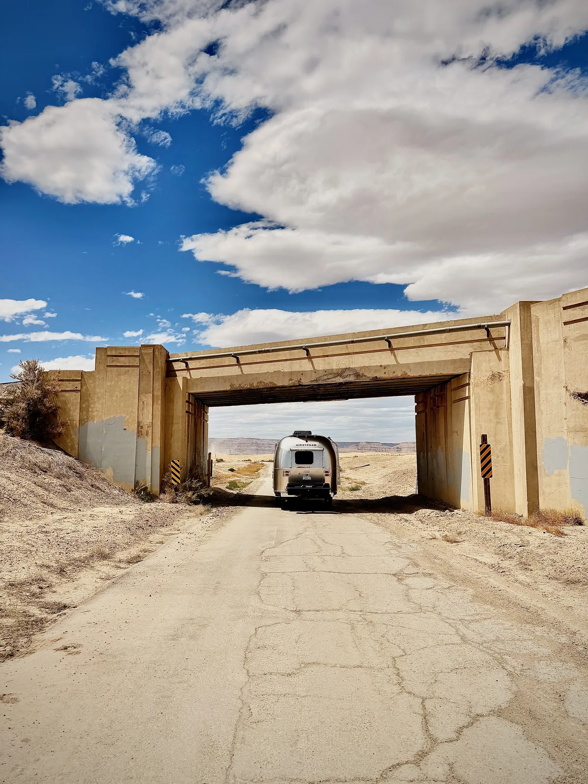 The Airstream and Rivian framed in the arch of an old desert highway underpass on a cracked dirt road under dramatic clouds