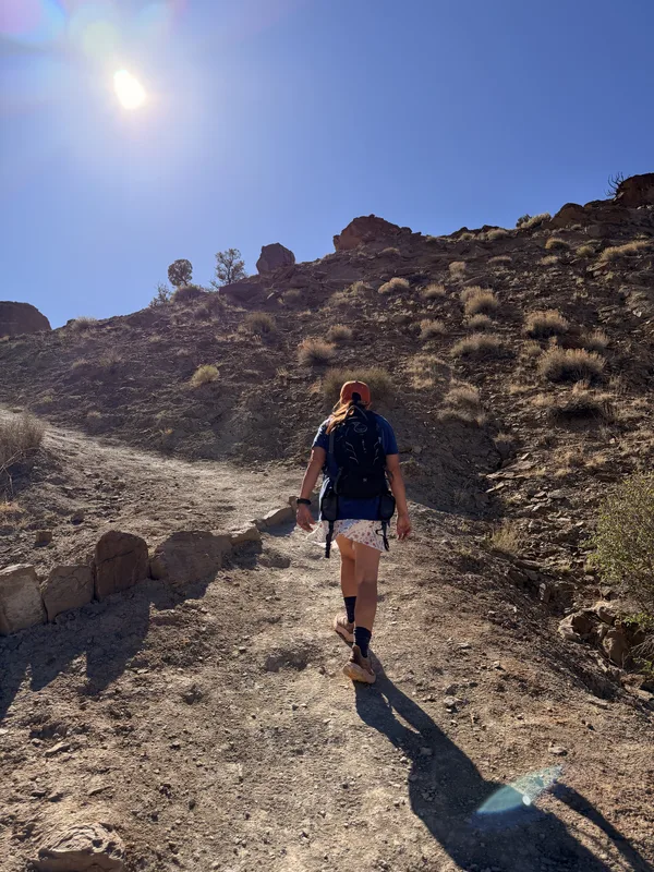 Diana hiking up the sunlit Chimney Rock trail in Capitol Reef, the dirt path climbing through sage toward the ridge