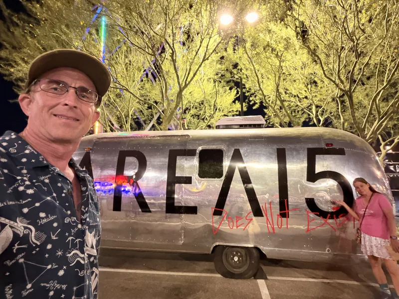 A polished Airstream trailer with AREA15 lettering on its flank parked at night outside the venue, Eric in the foreground