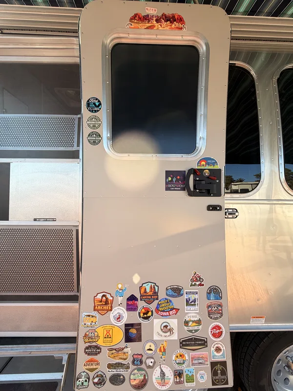 The entry door of the polished Airstream International "Smugglers Escape" covered in travel stickers from national parks and road-trip landmarks, photographed at the Oasis Las Vegas RV Resort
