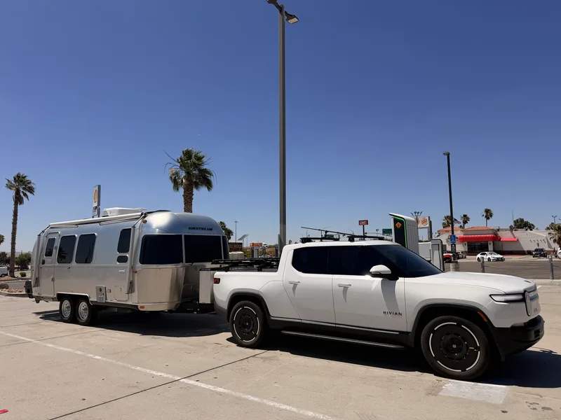 The polished silver Airstream and white Rivian R1T parked still hitched at a charging stall in a Barstow, California desert plaza under a deep blue sky, with palm trees and roadside signs behind them